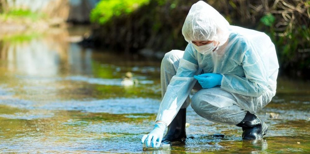 Image of a person in a chemical suit with mask collecting a water sample in a river.