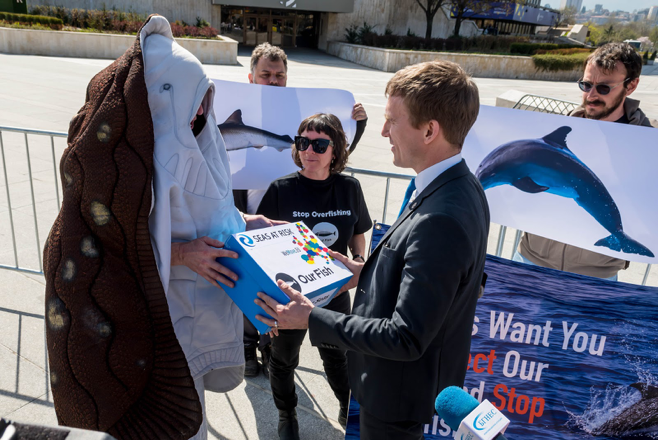 Ein Campaigner verkleidet als Rochen übergibt an einen Delegierten einen Karton gefüllt mit Unterschriften