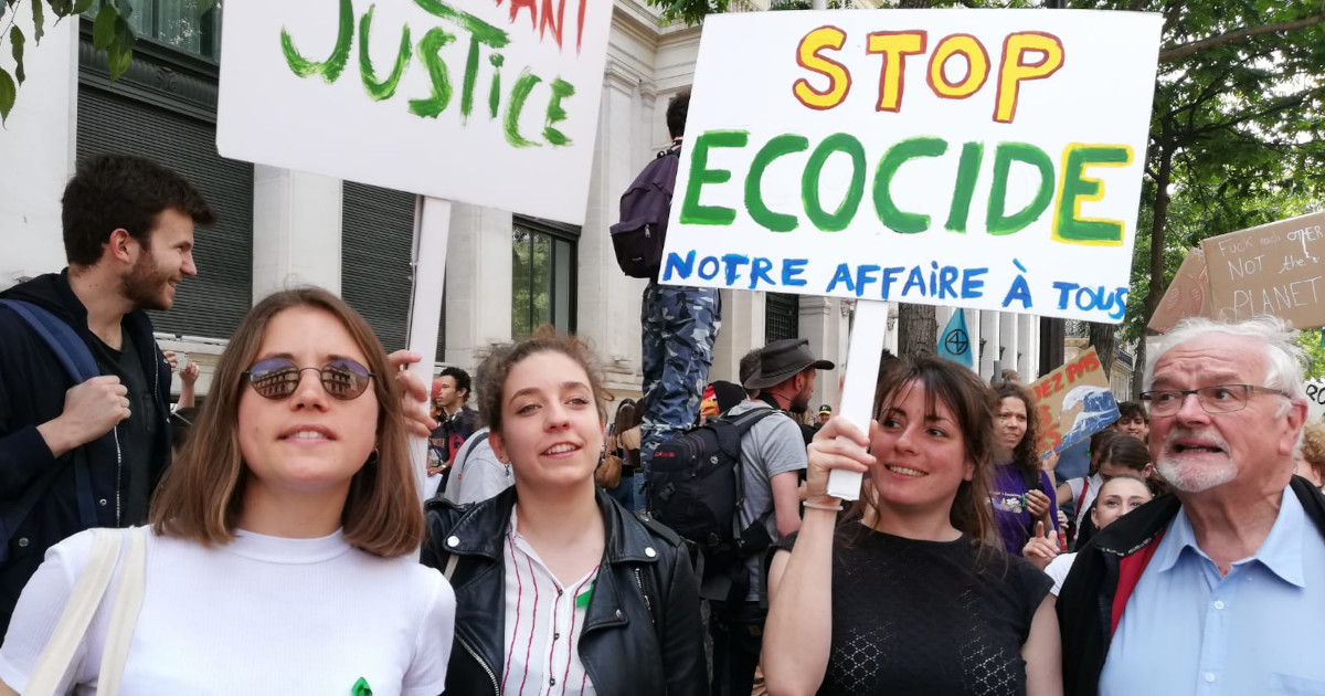 From left: three woman and one man stand outside, protest going on behind them, holding climate justice placard