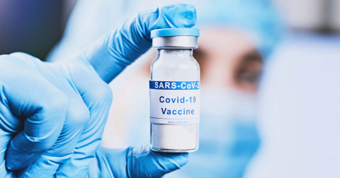 Eine Hand mit blauen Handschuhen hält ein Glasfläschchen mit der Aufschrift "Covid-19 Vaccine"